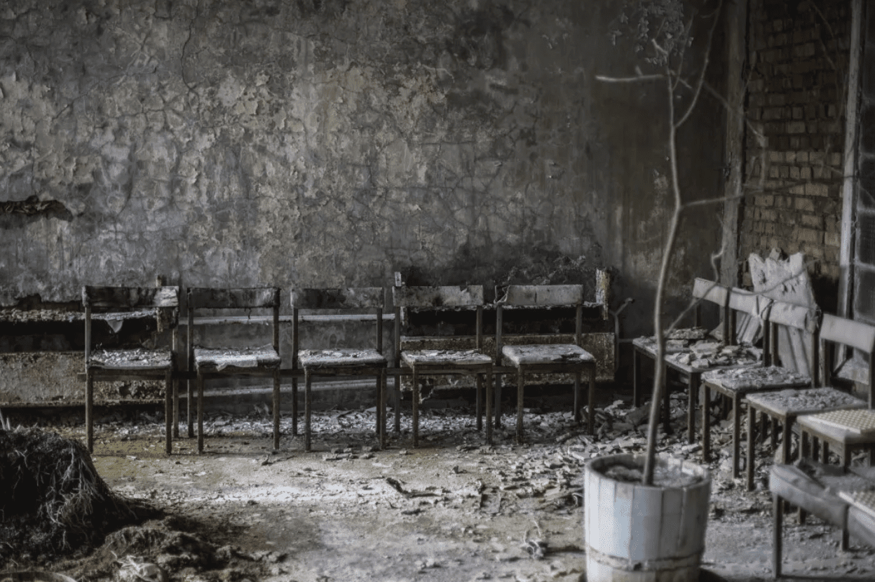 A black and white photo of what appears to be the corner of a burned out room.  One wall has peeling paint on damaged concrete and the other wall is exposed brick.  A line of 5 armless chairs stands against the concrete wall and another line of 5 armless chairs lines the exposed brick wall.  Ash and debris cover the floor and furniture.  A dead potted tree stands in the middle right of the room.  The image gives the feeling that people once gathered in this place of promise, but now this room is a shell of what it once was.
